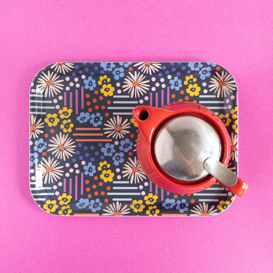 Colorful floral and geometric tray holding a red teapot on a pink background.