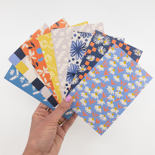 A hand holds 8 floral and geometric postcards in a fan arrangement.