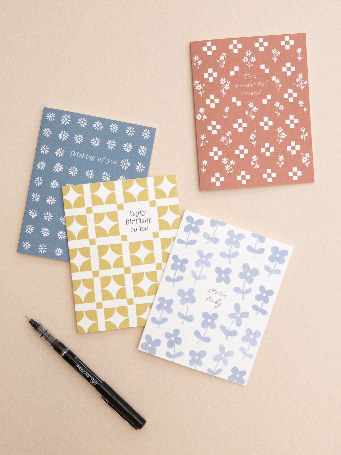 Four patterned greeting cards on a beige background with a pen.