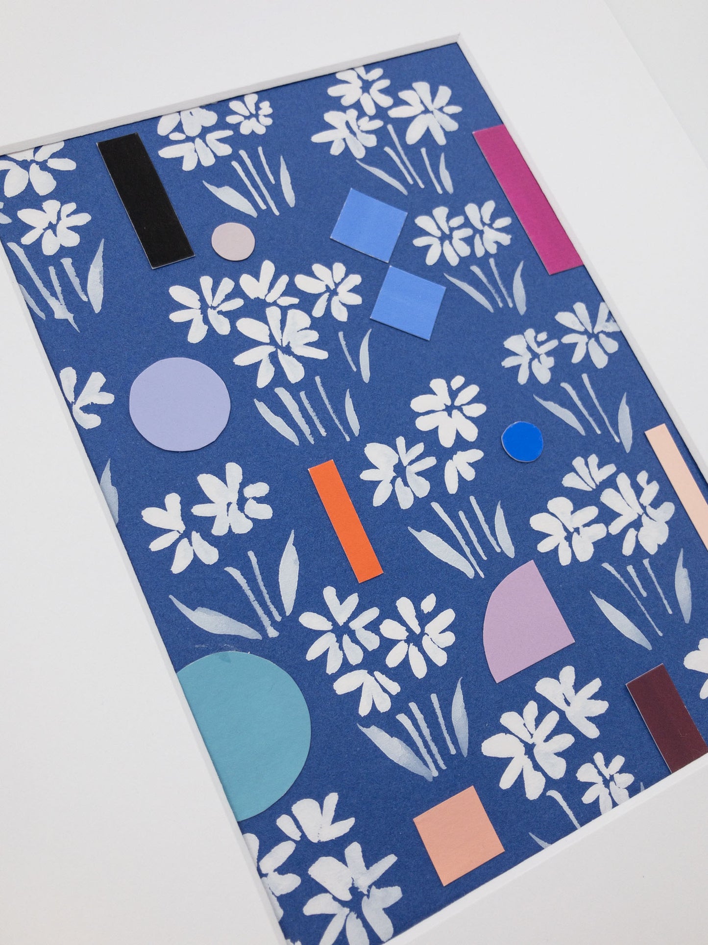Angled photo of artwork featuring hand-painted white florals on a blue background interspersed with colorful geometric shapes.