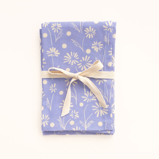 Folded periwinkle blue tea towel with a white floral pattern tied with a ribbon on a white background.