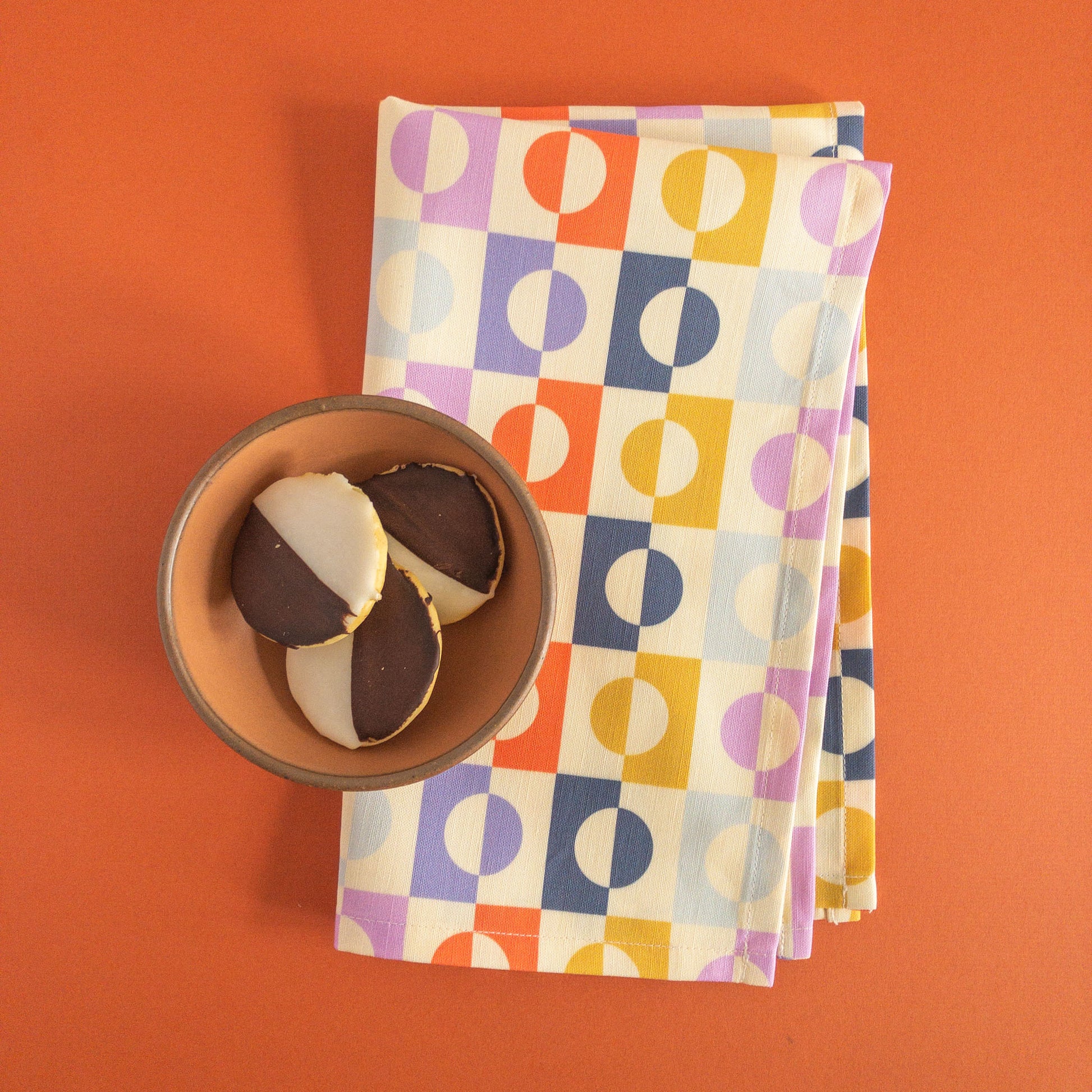 Folded tea towel with a colorful geometric pattern of circles and squares, styled on an orange background next to a bowl containing black-and-white cookies.