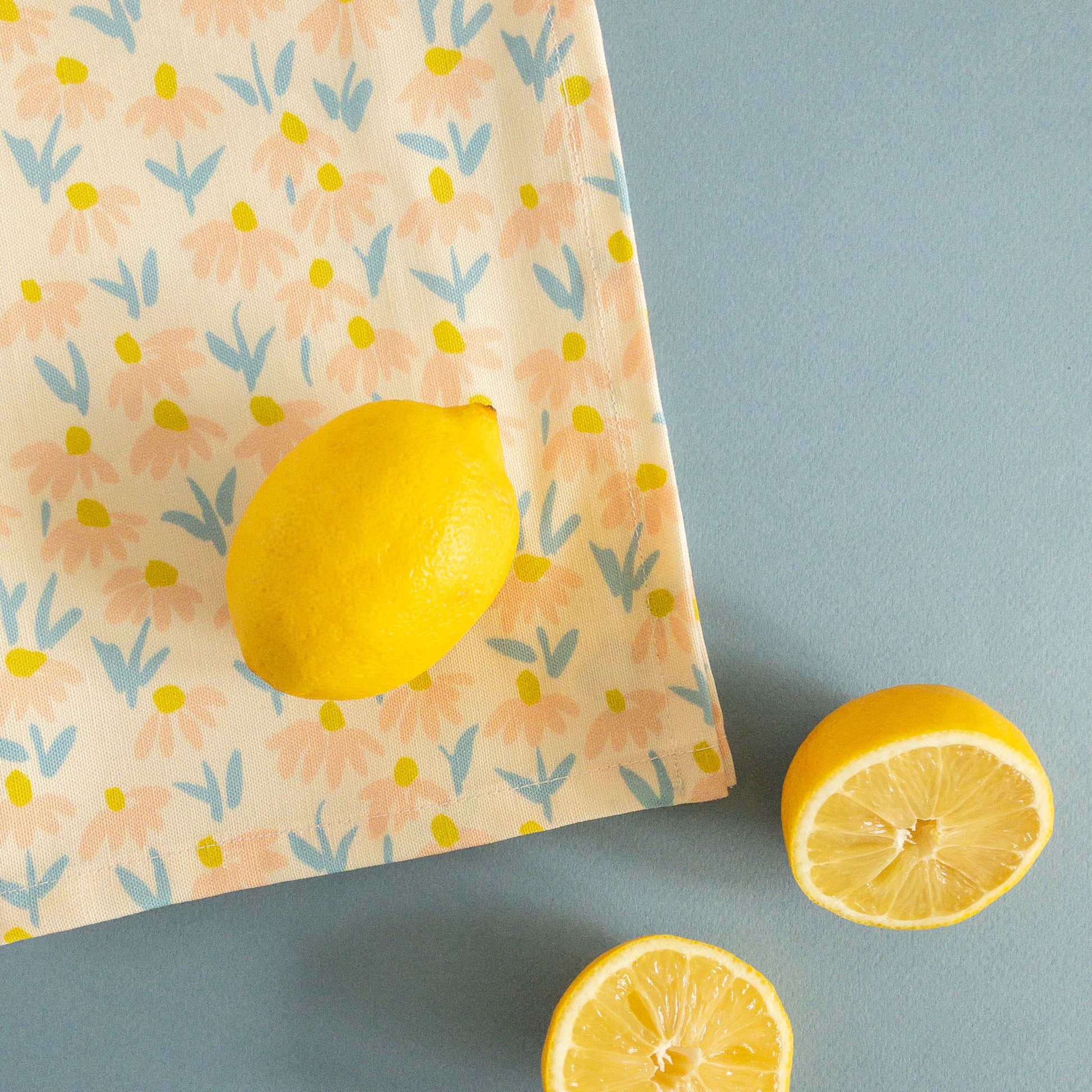 Floral tea towel featuring blue, peach, and yellow coneflowers styled on a blue background with lemons.