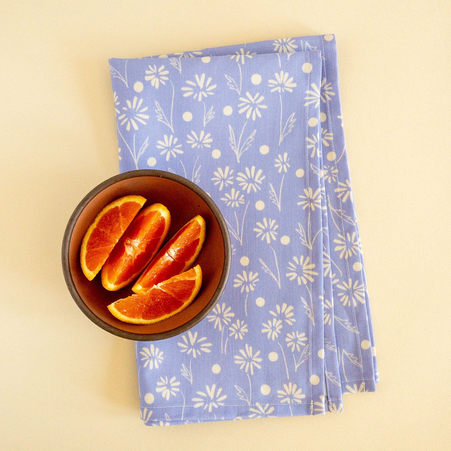 Folded blue floral tea towel styled on a cream background next to a bowl containing orange slices.