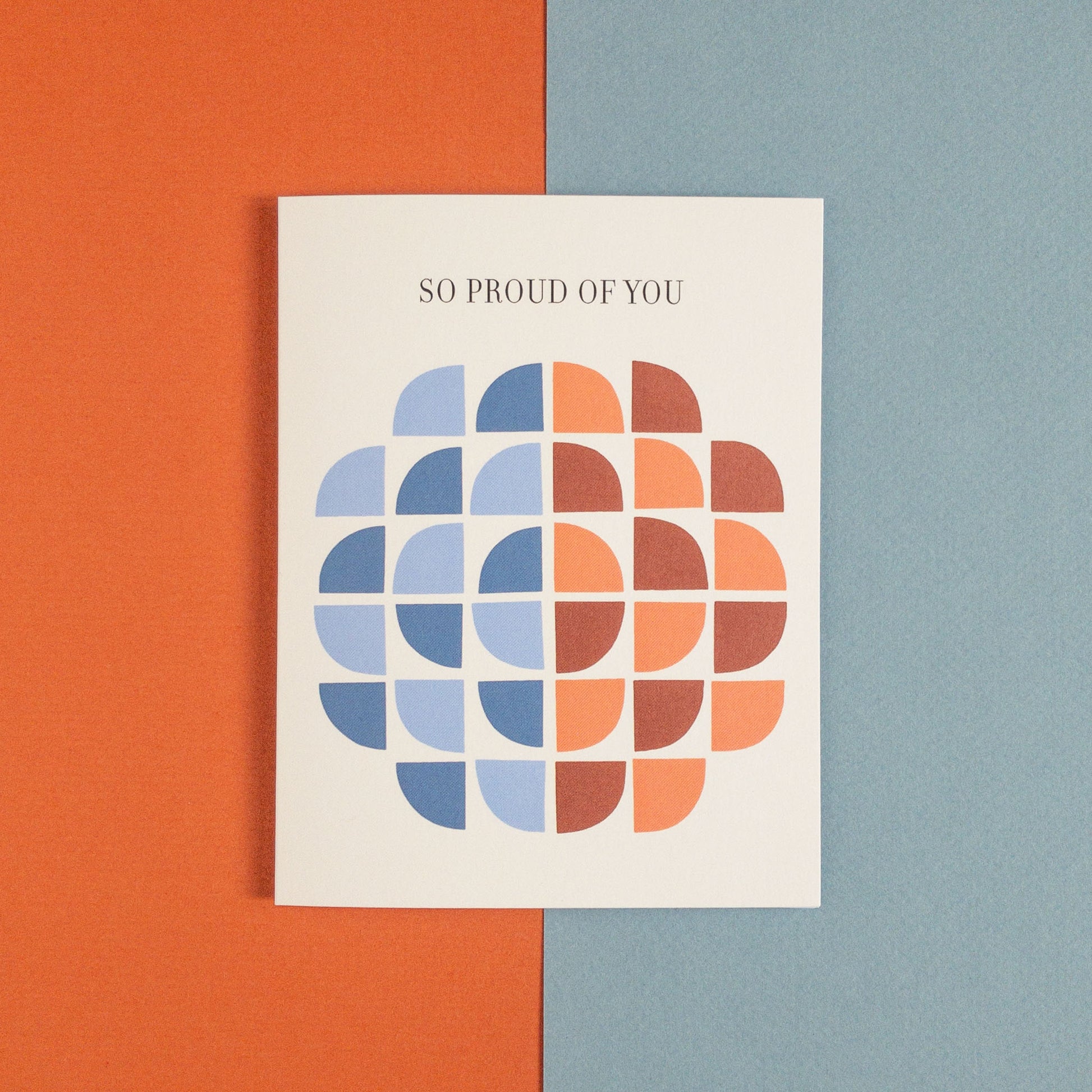 Geometric greeting card featuring quarter circles in blues and reds and the text "So Proud Of You" on a blue and orange background.