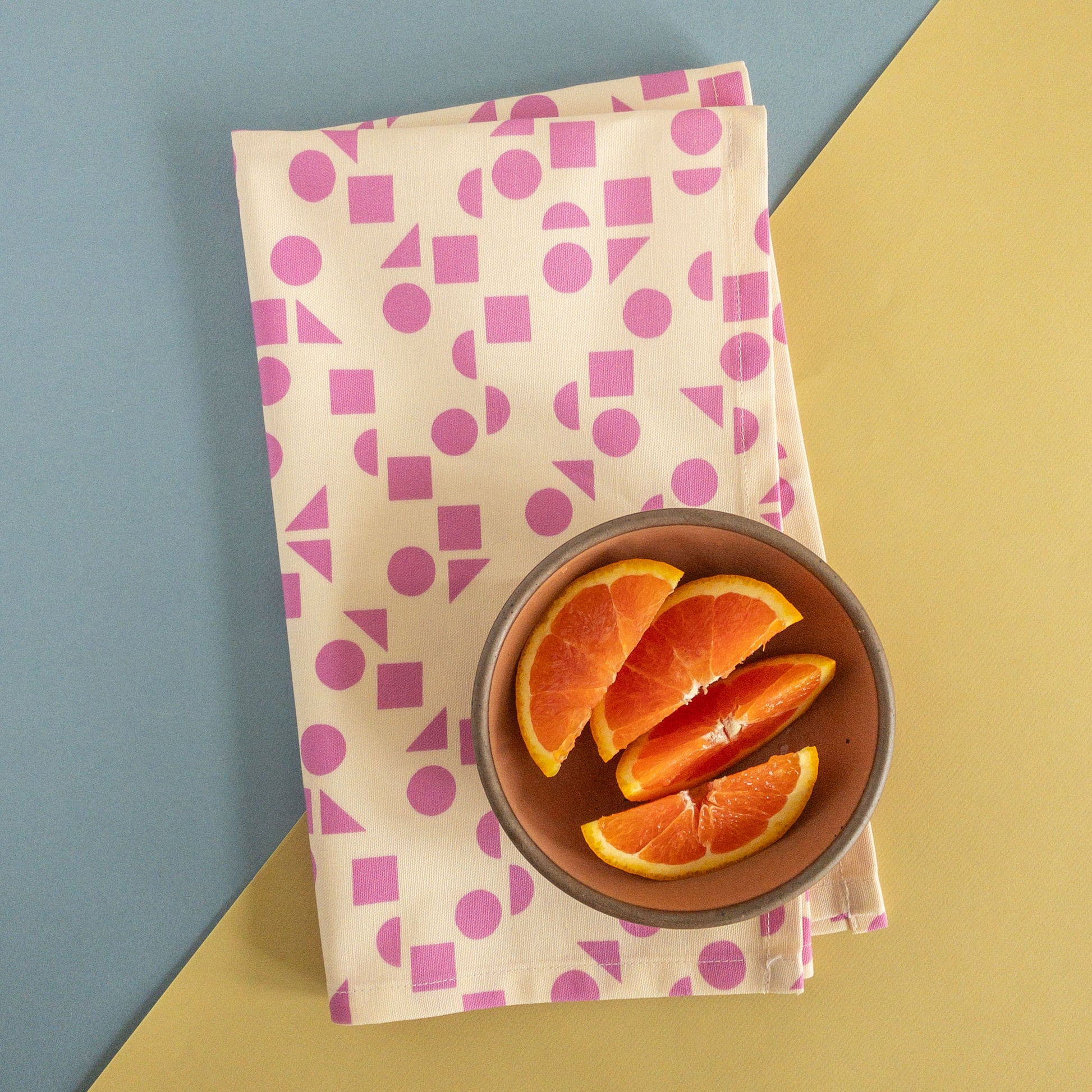 Folded geometric tea towel styled on a blue and yellow background next to a bowl containing orange slices.