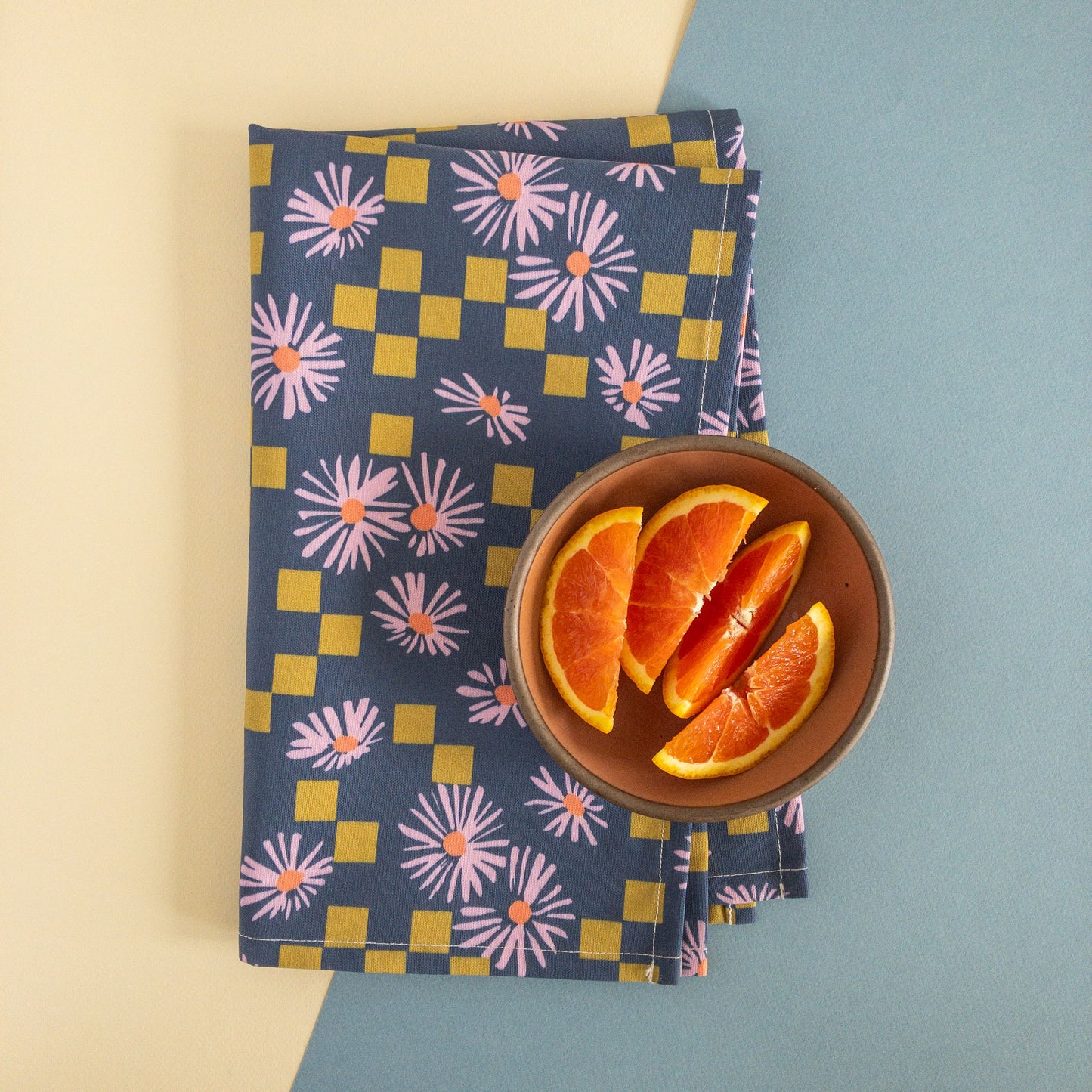 Modern floral tea towel featuring a pattern of purple daisies and olive squares, folded on a white and blue background, with a bowl of orange slices.