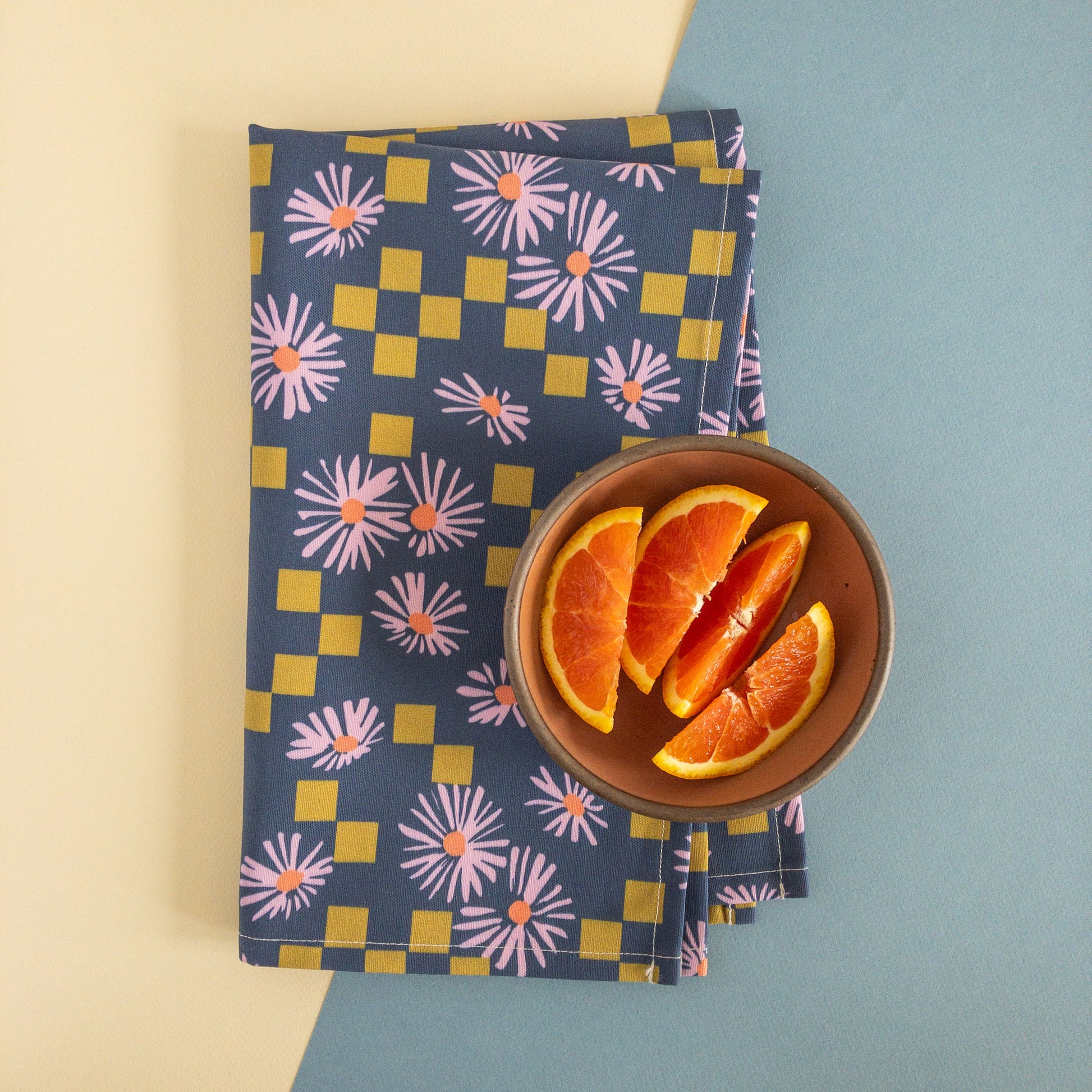 Modern floral tea towel featuring a pattern of purple daisies and olive squares, folded on a white and blue background, with a bowl of orange slices.