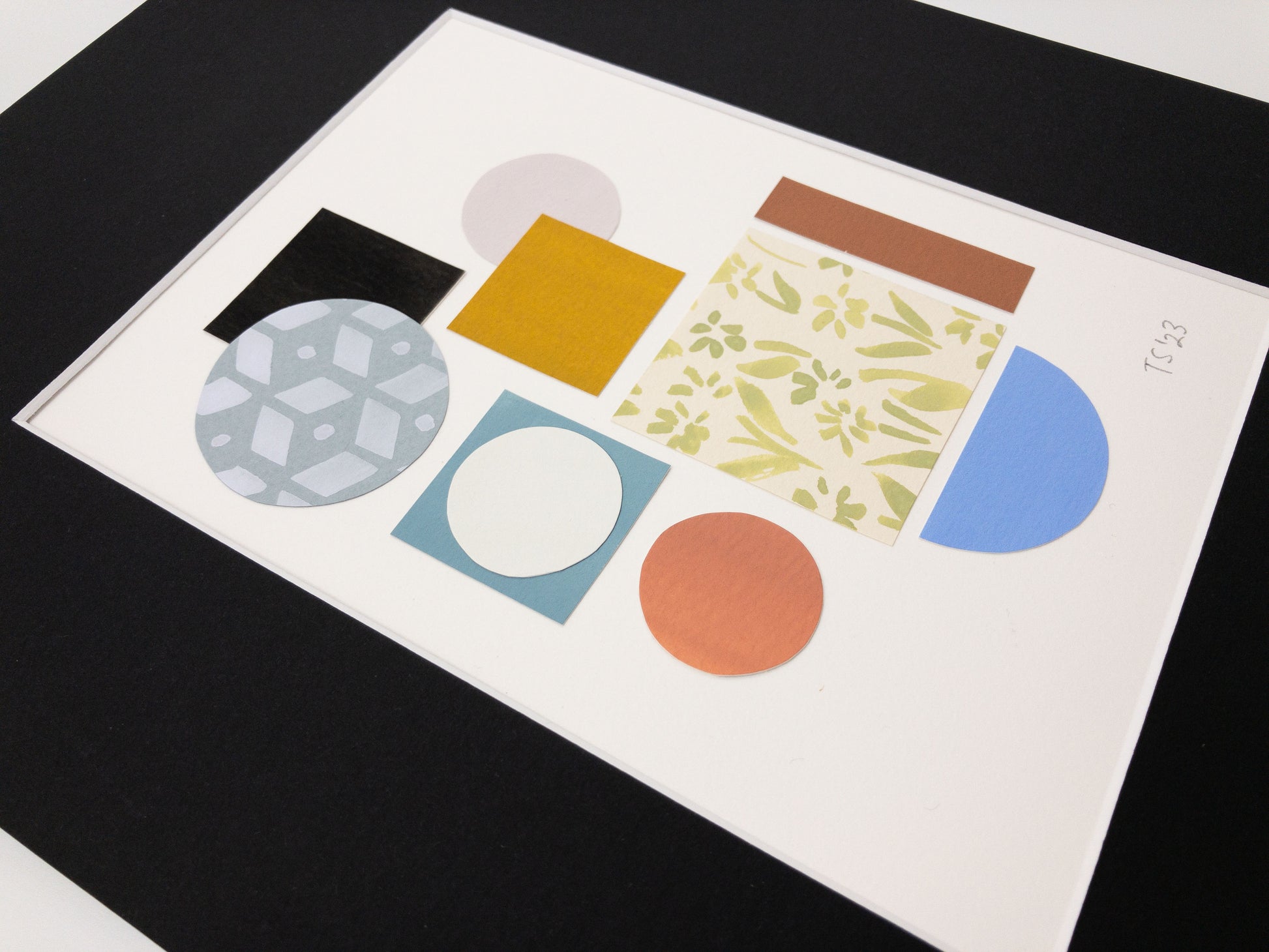Angled photo of a geometric collage featuring colorful circles, squares, and hand-painted pattern elements on a white background. The artwork is in a black mat.