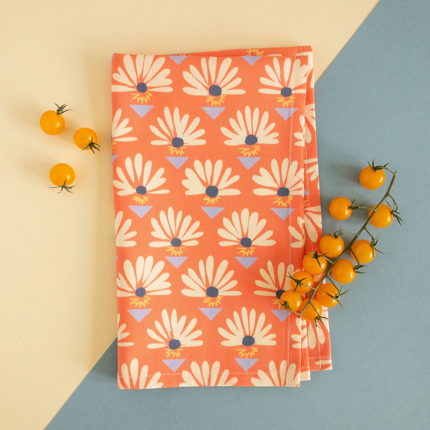 Folded red tea towel with daisies and blue triangles, styled with small yellow tomatoes.